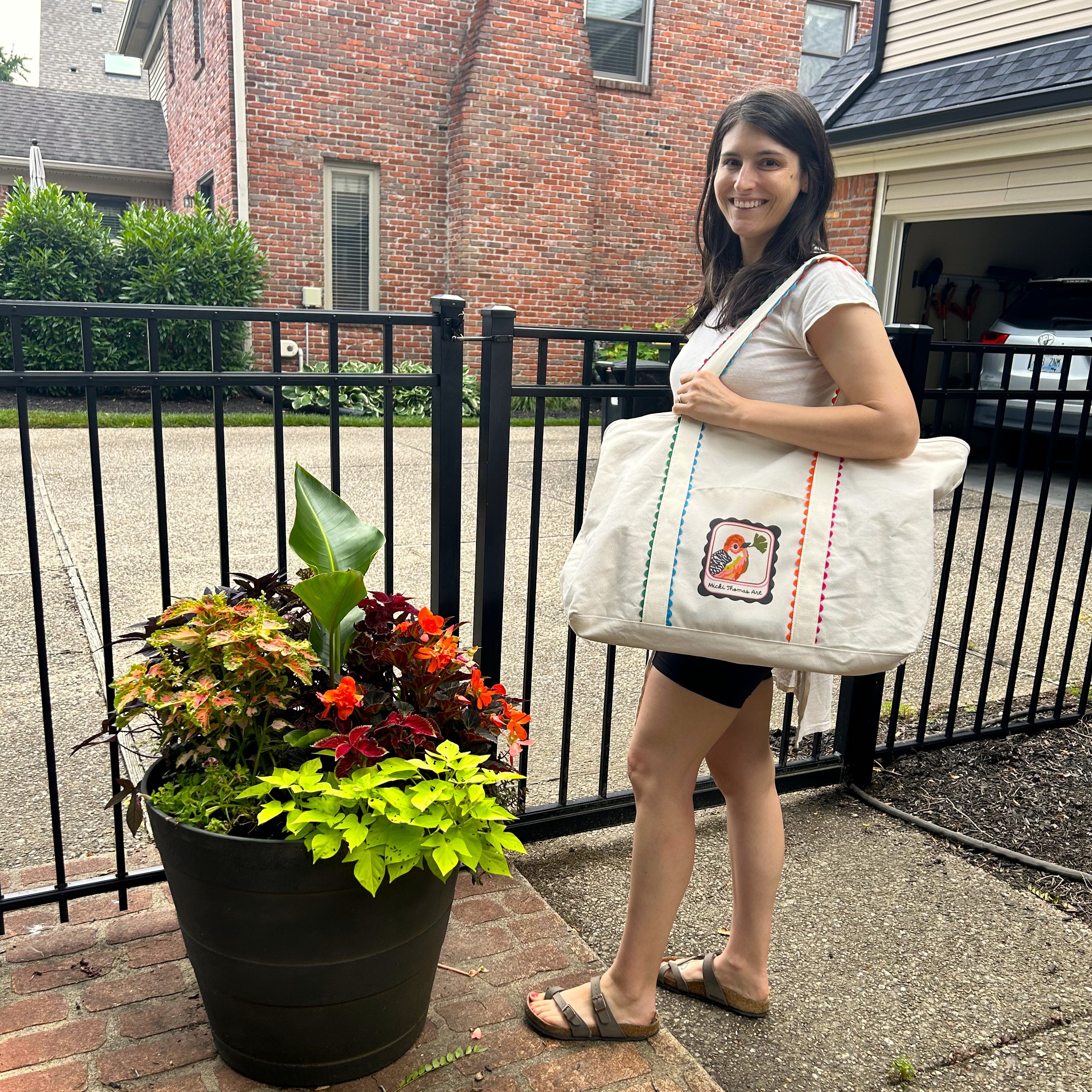 young brunette women holding a mahjong tote bag with a bird on it next to flowers